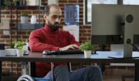 A man in a red sweater sits in a wheelchair working on a computer at a desk, possibly conducting accessibility testing. The background features a brick wall, a water cooler, and shelves with books and plants.
