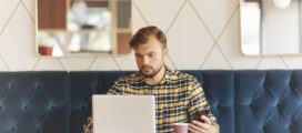 A man with a beard is sitting in a cafe, focused on his laptop while holding a smartphone in his other hand. There is a paper coffee cup on the table in front of him.