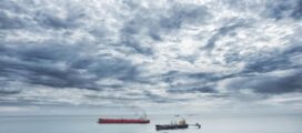 A cloudy sky over a calm sea with a red cargo ship and smaller vessels in the distance.