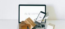 A small shopping cart with a smartphone and credit card sits on a laptop, surrounded by small packages. The laptop screen displays the text "ONLINE SHOPPING.