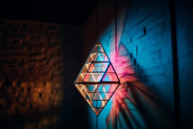 Geometric metal sculpture on a textured wall casts colorful shadows with blue and orange lighting.