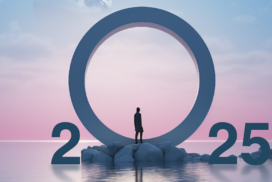 A person stands on rocks in front of a large ring between the numbers 2025, symbolizing the dawn of tech predictions, set against a pink and blue sky over water.