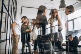 A group of six people stand and talk in a modern office, some discussing software development methodologies while others use phones near large windows and glass walls.