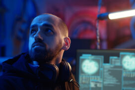A man with a beard and headphones around his neck looks to the side in a dimly lit room with computer monitors displaying data in the background.