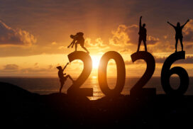 Four people pose on large "2026" numerals at sunset by the sea, silhouetted against an orange sky—capturing a moment of anticipation for 2025 tech predictions as the sun meets the horizon.