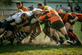 Two rugby teams, one in white and the other in orange, engage in a scrum on a grassy field during a match, moments before a potential scrum failure.