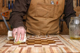 A person in a brown apron wipes oil onto a wooden chessboard in a workshop, with tools and AI tools on the wall, and a jar of oil on the table.