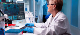 Scientist in a lab coat and gloves working at a computer in a laboratory, with scientific equipment and another researcher in the background.