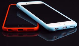 On a black background two smart phones, one on top of the other crossed, the top with a white case, the bottom with a red case