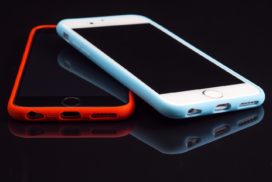 On a black background two smart phones, one on top of the other crossed, the top with a white case, the bottom with a red case