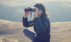 On a windswept hill a squatting woman peers through binoculars.