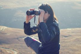 On a windswept hill a squatting woman peers through binoculars.
