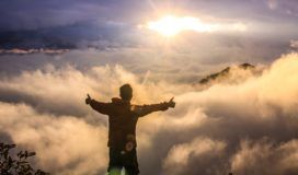 man looking down on clouds standing on a mountain with his thumbs up