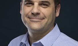 A man in a blue shirt smiling in front of a black background.