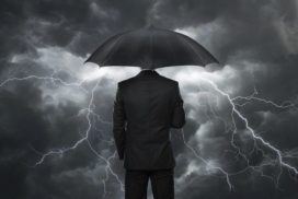 A businessman in a black suit holding a black umbrella stands in front of a gray-clouded sky while lightning explodes all around.