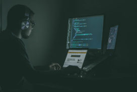 A young man with a beard wearing headphones and glasses works on a laptop while looking at code on a secondary monitor.