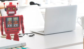 A red robot sitting on a desk next to a laptop.