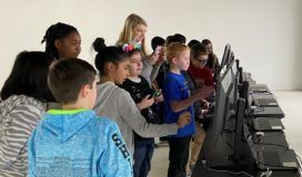 A group of children standing in front of a group of computers.