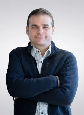 A man with brown hair, wearing a dark jacket over a light shirt, stands with arms crossed and smiles slightly against a plain light background.