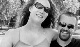 An adult woman wearing sunglasses and a halter top sits in the lap of an adult male wearing sunglasses.