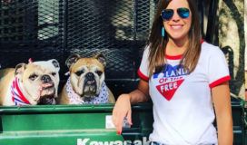An adult woman wearing sunglasses and a white t-shirt stands next to two bulldogs in a truck bed.