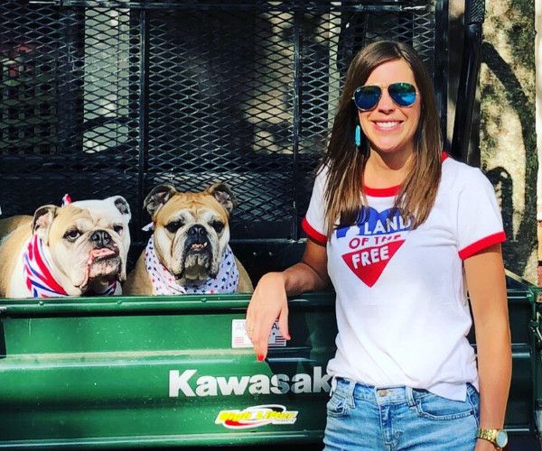 An adult woman wearing sunglasses and a white t-shirt stands next to two bulldogs in a truck bed.