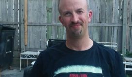 An adult male wearing a t-shirt that reads "Revolution Brewing" sits outdoors in front of a wooden fence.
