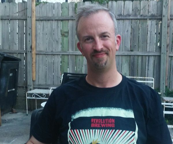 An adult male wearing a t-shirt that reads "Revolution Brewing" sits outdoors in front of a wooden fence.