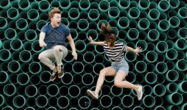 Two individuals jumping in front of a group of green pipes.