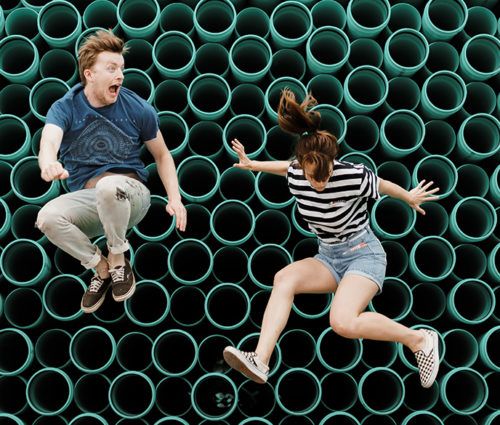 Two individuals jumping in front of a group of green pipes.