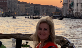 An adult woman standing in a motor boat on a canal flanked by buildings as the sun sets.