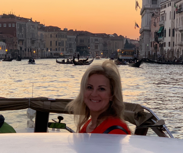 An adult woman standing in a motor boat on a canal flanked by buildings as the sun sets.