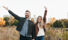 Adult male and adult female holding hands and raising their other arms while standing on a sidewalk flanked by a field.