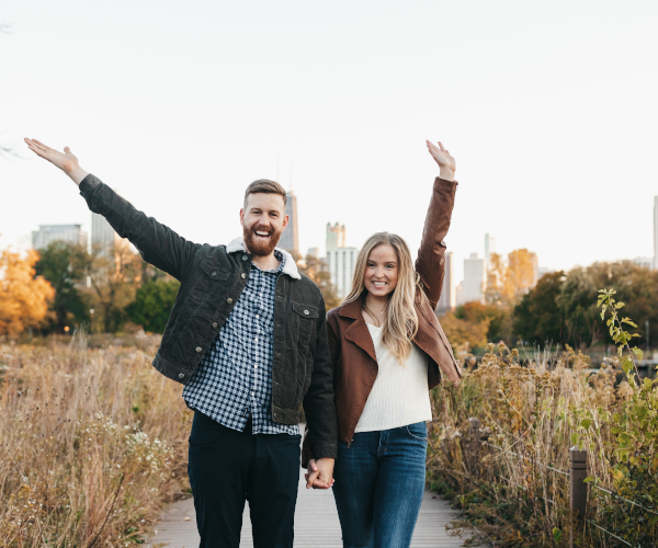 Adult male and adult female holding hands and raising their other arms while standing on a sidewalk flanked by a field.