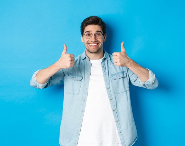 Attractive young man wearing glasses and casual clothes, showing thumbs up in approval, like something, standing against blue background.