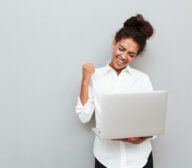 Photo of young african cheerful business woman standing over grey wall with laptop computer. Looking aside make winner gesture.
