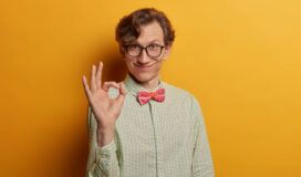 Funny smiling male geek shows ring gesture with fingers, says good job, satisfied with suggestion, wears spectacles and formal shirt, looks gladfully at camera, isolated on yellow background