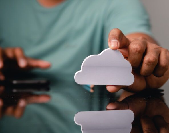 A person holds a white cloud-shaped cutout, symbolizing modern technology predictions, while another hand interacts with a smartphone on a reflective surface.