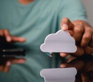 A person holds a white cloud-shaped cutout, symbolizing modern technology predictions, while another hand interacts with a smartphone on a reflective surface.