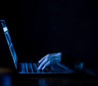 A hand types on a laptop keyboard in a dark room, the screen's blue light casting shadows as technology predictions unfold on the glowing display.