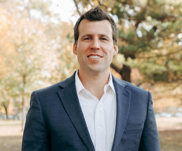 Adult male wearing a white collared shirt and a gray sport coat stands in a park.