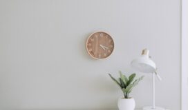 Against a neutral colored wall, a simple plant and lamp stand on a white table while a clock hangs on the wall.