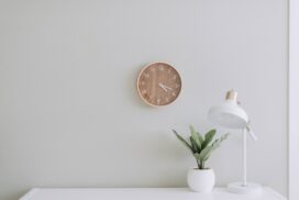 Against a neutral colored wall, a simple plant and lamp stand on a white table while a clock hangs on the wall.