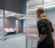 Man walking towards the glass door of an office labeled "admissions" in a modern technology firm, with a seating area visible in the background.