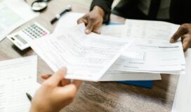 Two people sitting at a desk exchanging documents. Various papers, a calculator, and pens are spread out on the wooden table, hinting at an intricate discussion possibly involving machine learning applications.