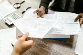 Two people sitting at a desk exchanging documents. Various papers, a calculator, and pens are spread out on the wooden table, hinting at an intricate discussion possibly involving machine learning applications.