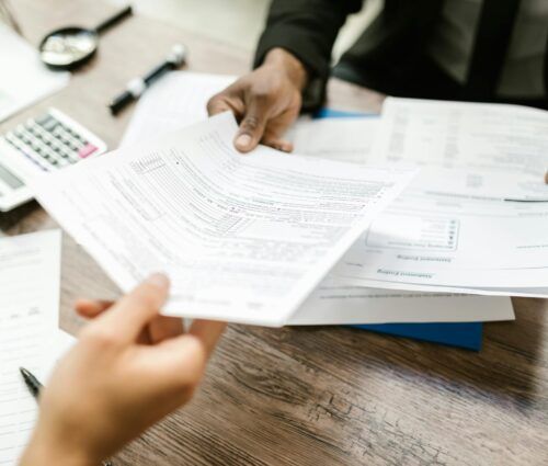 Two people sitting at a desk exchanging documents. Various papers, a calculator, and pens are spread out on the wooden table, hinting at an intricate discussion possibly involving machine learning applications.