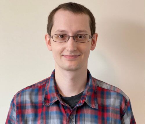 A smiling person with short hair and glasses, donning a red and blue plaid shirt, stands against a beige background, embodying the essence of test optimization.