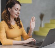 A woman in a yellow shirt sits at a table in front of a laptop, raising her hand as if waving or gesturing during an online meeting for accessibility testing.