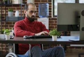 A man in a red sweater sits in a wheelchair working on a computer at a desk, possibly conducting accessibility testing. The background features a brick wall, a water cooler, and shelves with books and plants.